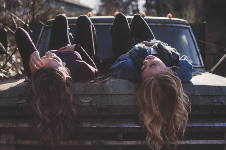 dwie młode kobiety smiejące się, rozmawiające i leżące na masce auta
