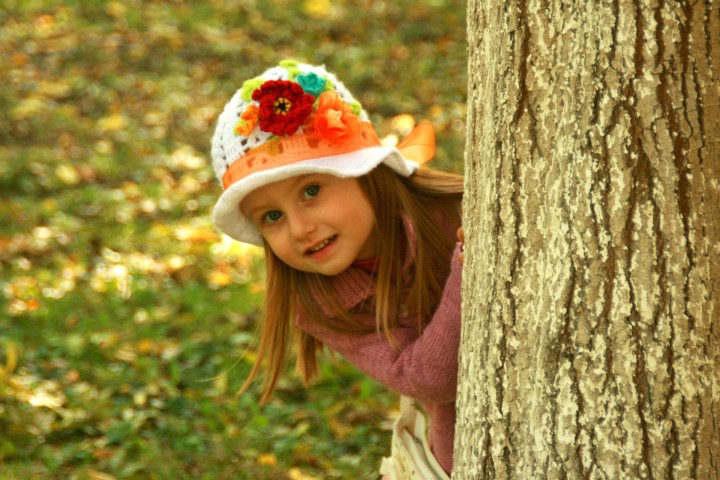mała dziewczynka w kolorowym kapeluszu chowająca się za pniem drzewa